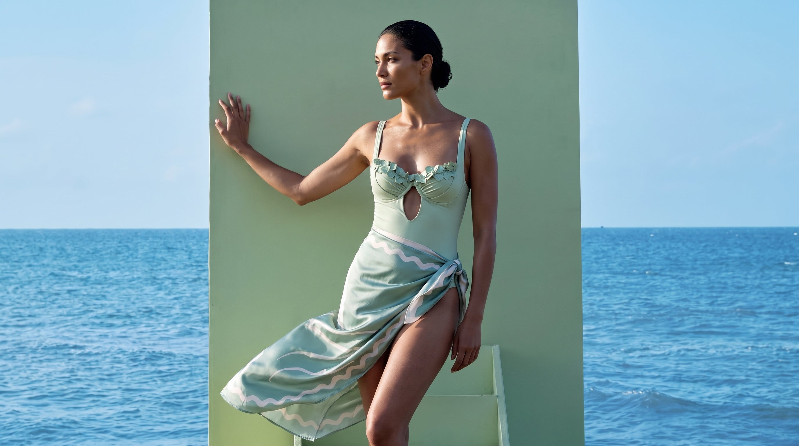 Mint one-piece and pareo on the green wall above the sea