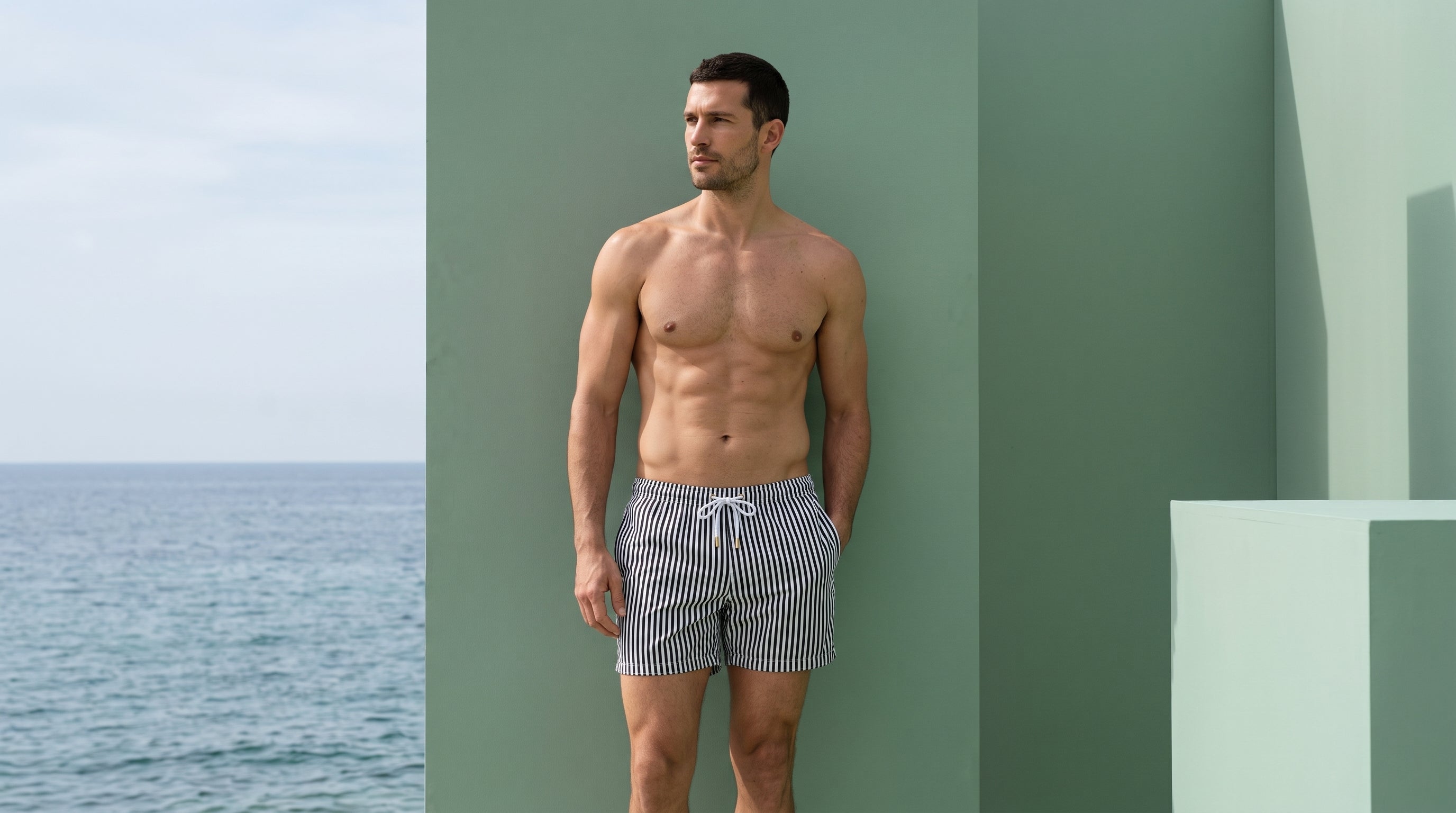 Man in striped ANCORA swim trunks against sage-green architecture by the sea